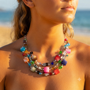A close-up lifestyle shot of a woman on a beach wearing a multi-strand statement necklace made of colorful translucent square beads.