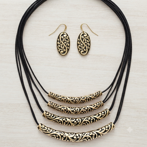 A jewelry set featuring a multi-strand black cord necklace with ornate gold filigree bar beads and matching oval drop earrings, displayed on a light wood background.