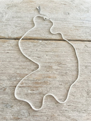 A delicate silver-toned beaded necklace with a lobster clasp, laid flat on a light-colored, rustic wooden surface.