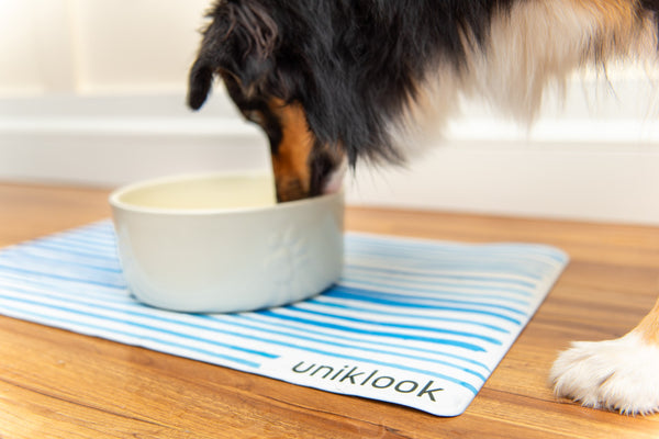 A pet food mat with an abstract blue line design, featuring the brand name 'Uniklook', placed on a wooden floor with a dog eating from a bowl on top of the mat.