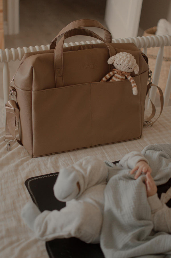 Brown diaper bag with a sheep toy on a bed, with a person partially visible.