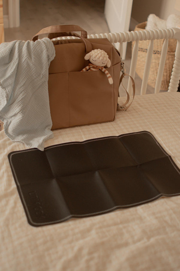 Brown diaper bag with a plush toy and a matching brown changing mat on a bed.