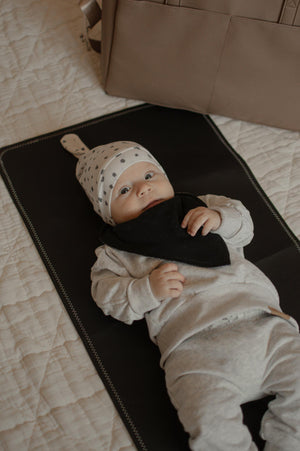 Baby lying on a black mat wearing a white outfit and hat with black polka dots.