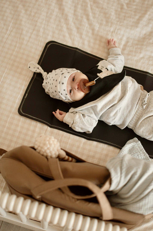 Baby lying on a black mat with a pacifier, surrounded by bags and clothes on a beige surface.