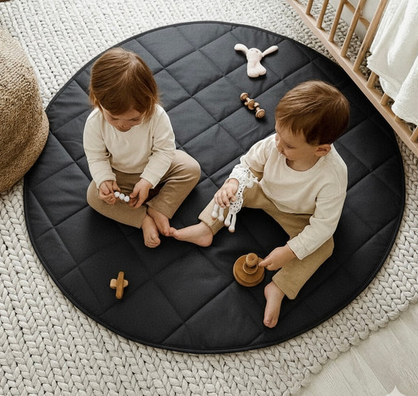 Two children playing on a round black mat with toys in a nursery setting.
