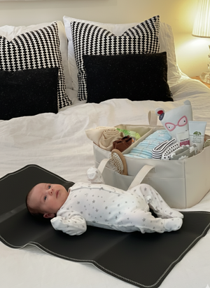 Baby lying on a black mat with a basket of items on a bed.