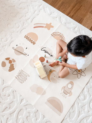 A child sitting on a beige play mat with a cosmic and terracotta design, including various icons and the word 'Cosmo'.