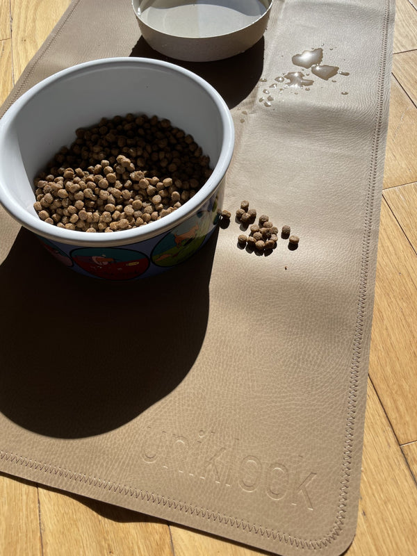 A tan pet bowl mat with a gray border, placed on a wooden floor, with a bowl of pet food and a dish with water on top of it placed on the mat.
