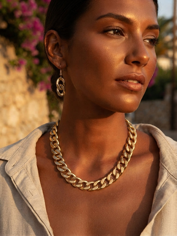 A close-up of a woman wearing a chunky gold Cuban link chain necklace and matching gold hoop earrings, photographed outdoors in warm golden hour light.