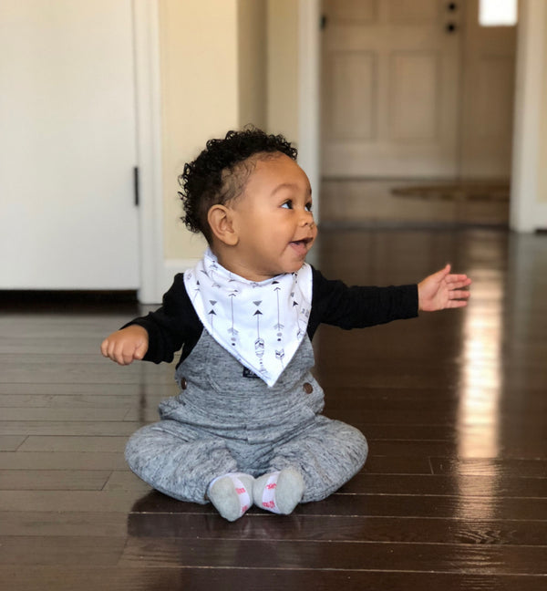 A baby wearing a black and gray bandana bib with a pattern, sitting on a floor.