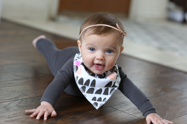 A baby lying on a floor wearing a gray long-sleeve top and a black and white bandana bib with a bow tie.
